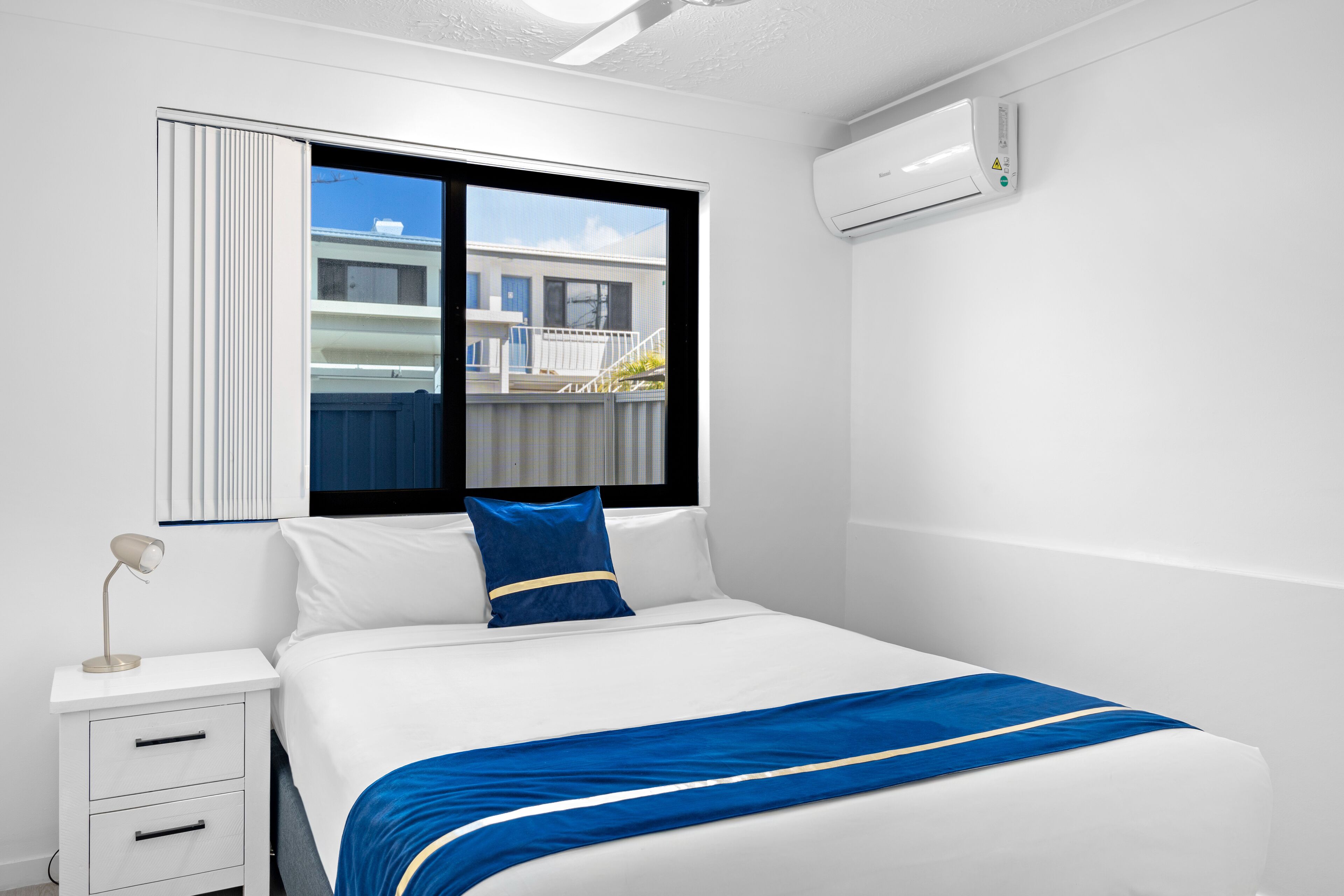 Ein modernes Hotelzimmer mit einem Bett, einem Nachttisch, einer Lampe, einem Fenster mit Blick auf Gebäude und einer Klimaanlage.