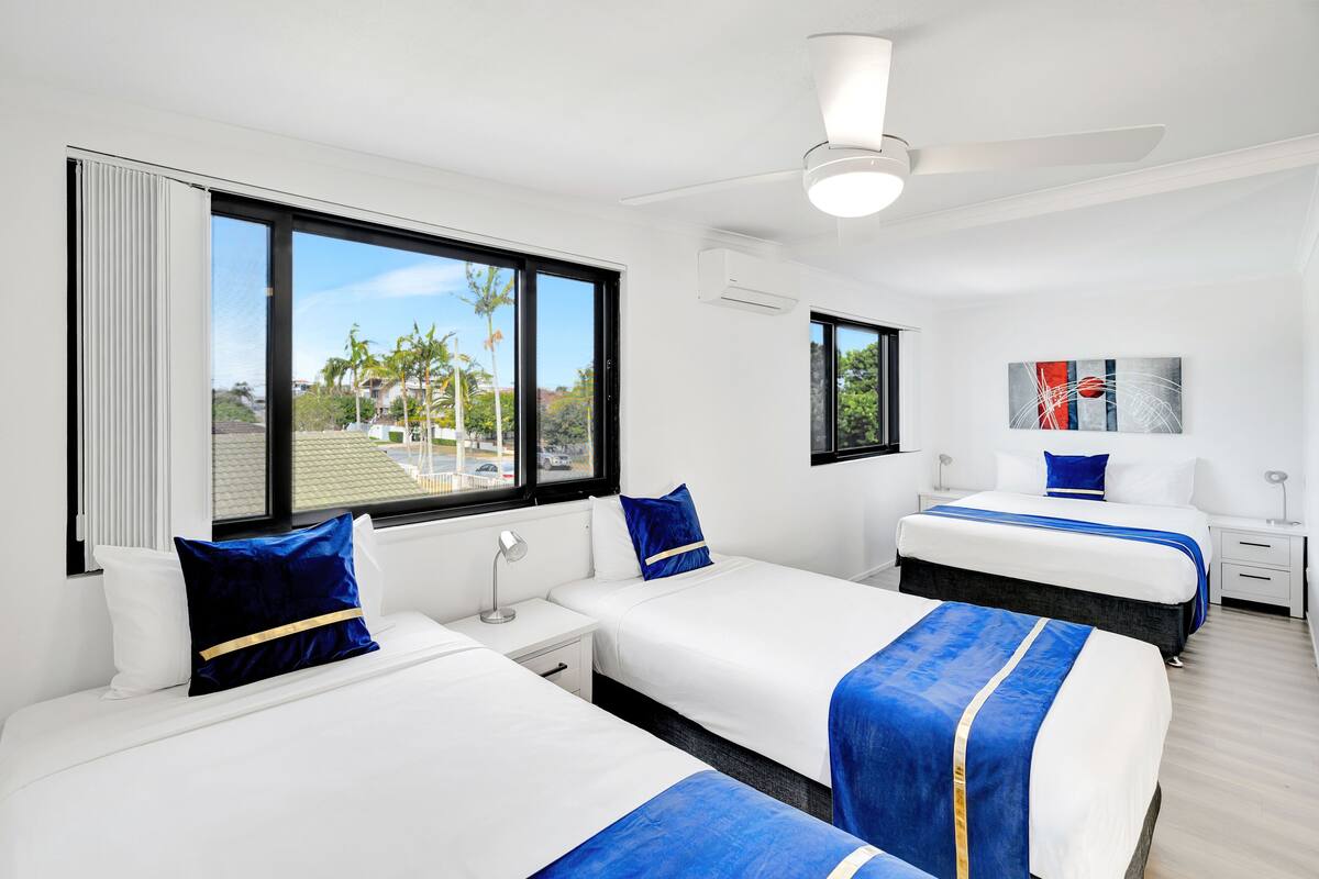 A hotel room with two beds, a ceiling fan, and large windows offering a view of palm trees and buildings.