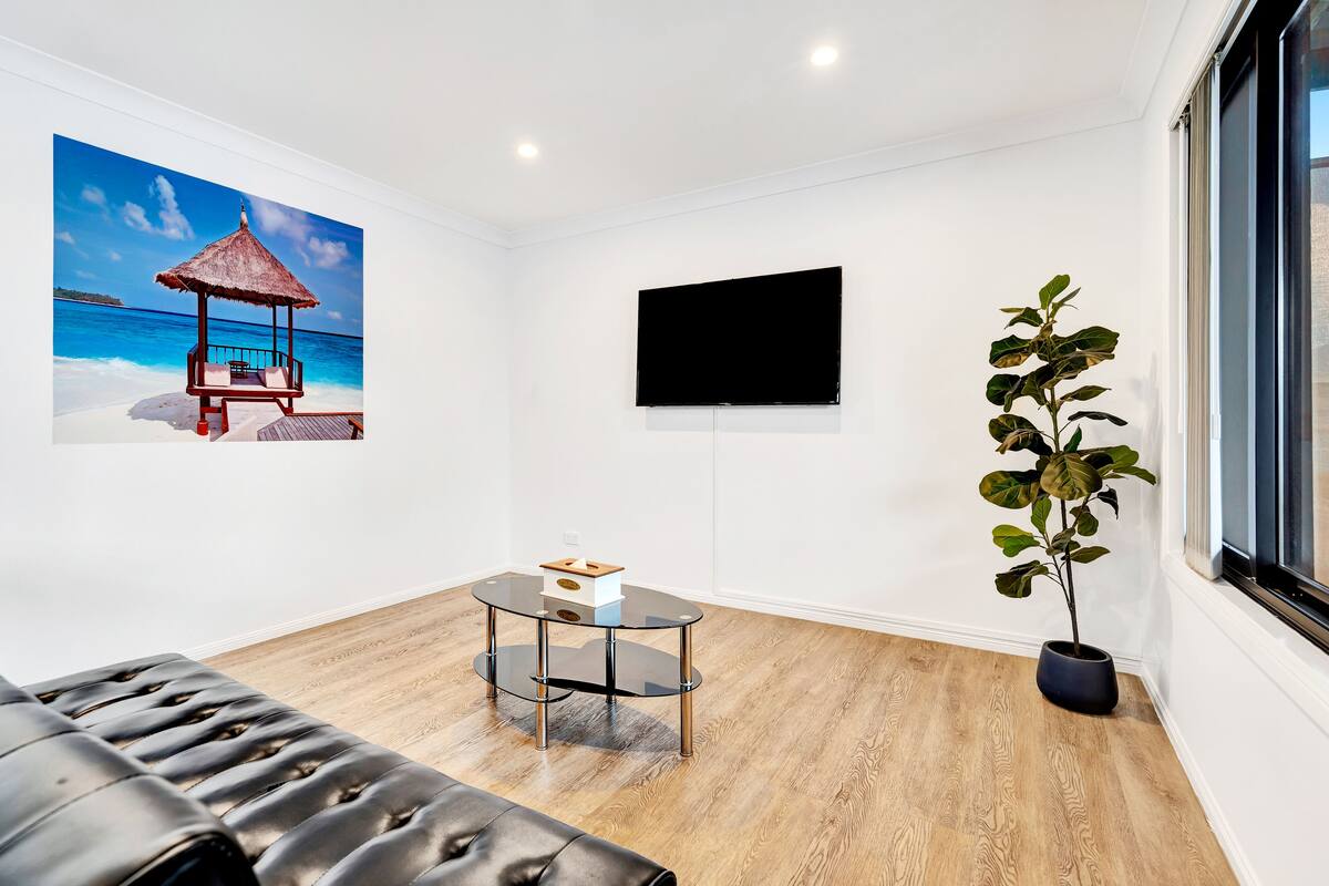 A modern room with a wooden floor, a glass table with a box, a black leather sofa, a TV on the wall, and a potted plant.