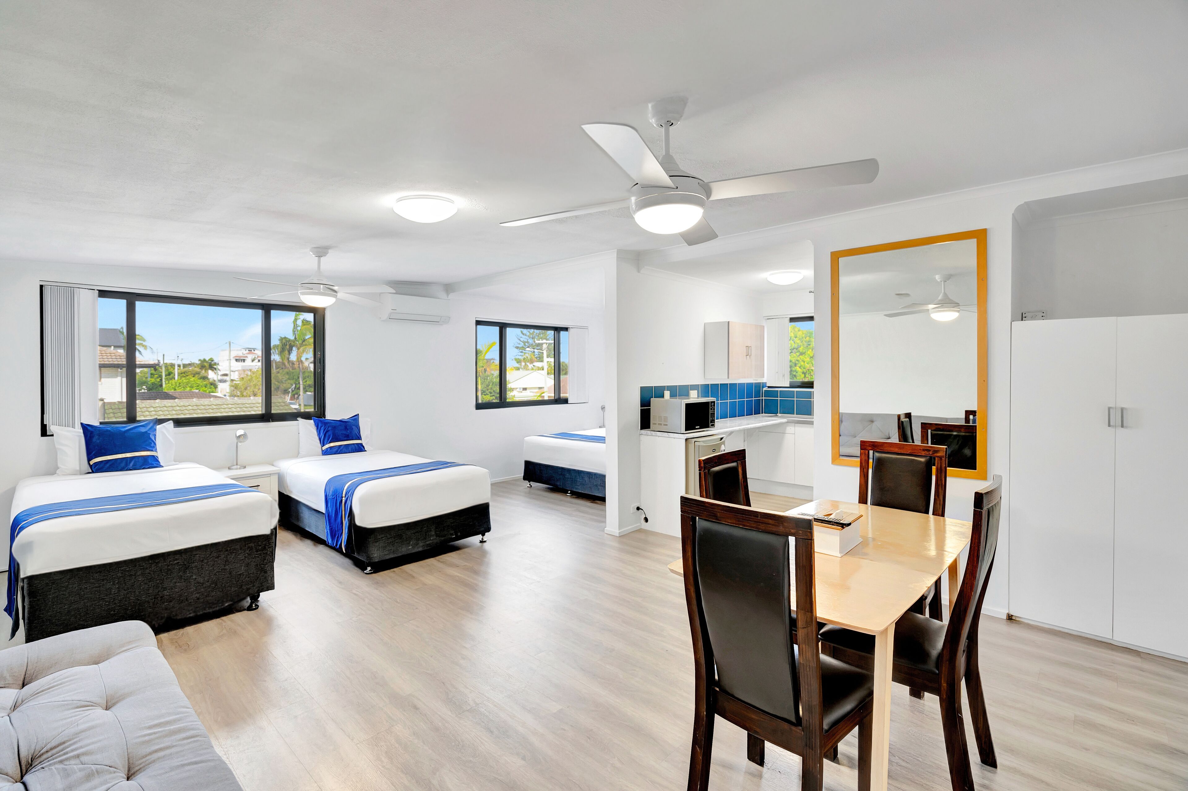 Una habitación de hotel moderna con dos camas, una mesa de comedor, sillas, un ventilador de techo y grandes ventanales que ofrecen vistas al exterior.