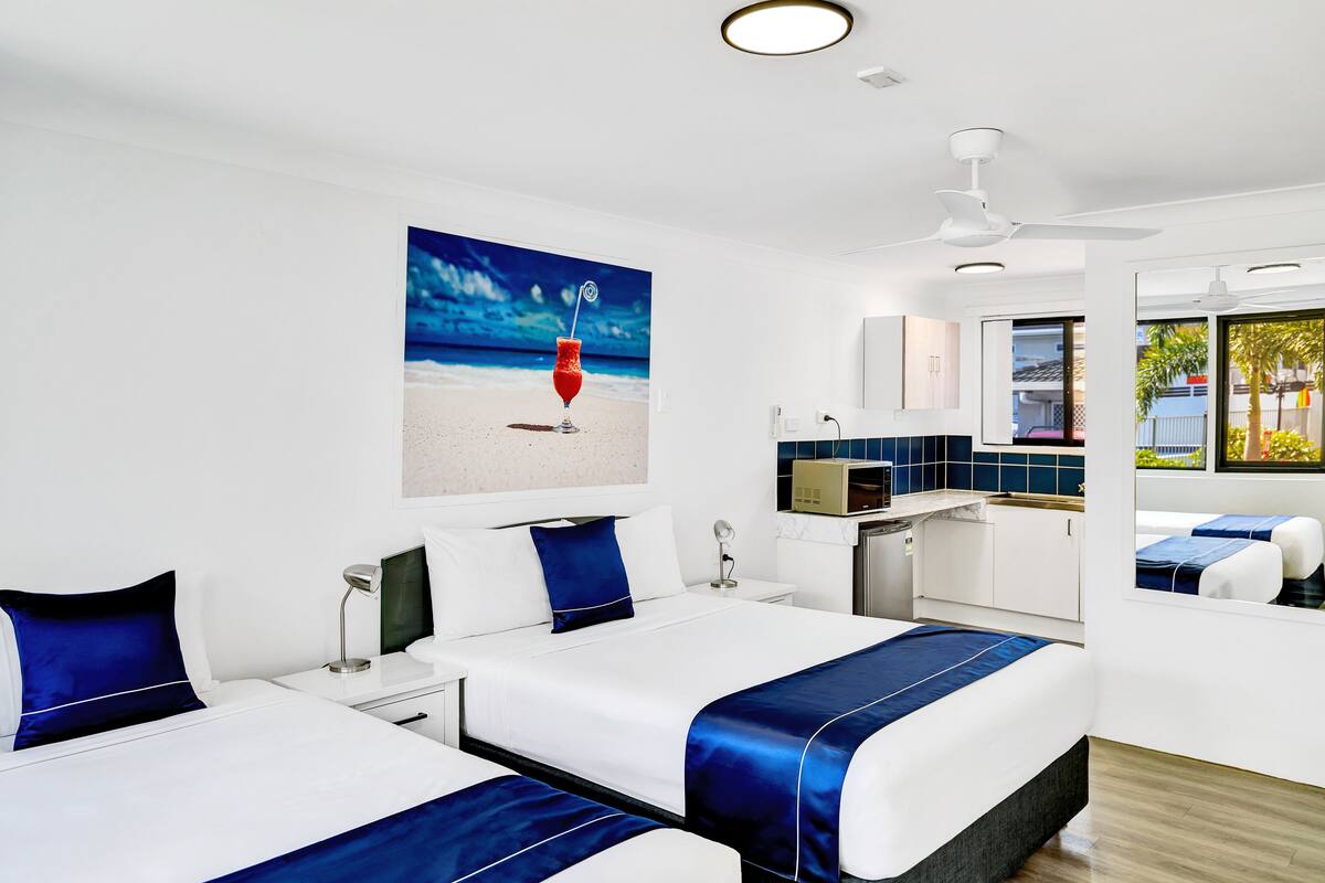 A hotel room with two beds, a painting of a beach scene, a ceiling fan, and a view of the outside.