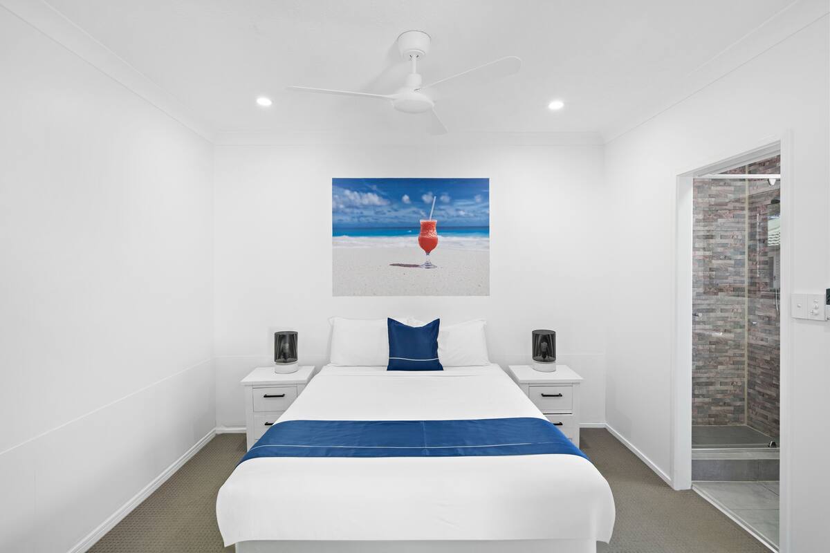A modern hotel room with a bed, white walls, a ceiling fan, a beach-themed artwork, a brick shower enclosure, and a nightstand with a lamp.