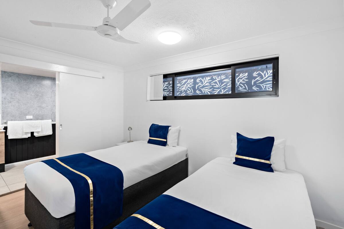 A hotel room with two beds, a ceiling fan, a window with leaf-patterned blinds, a towel rack, and a small bathroom area.