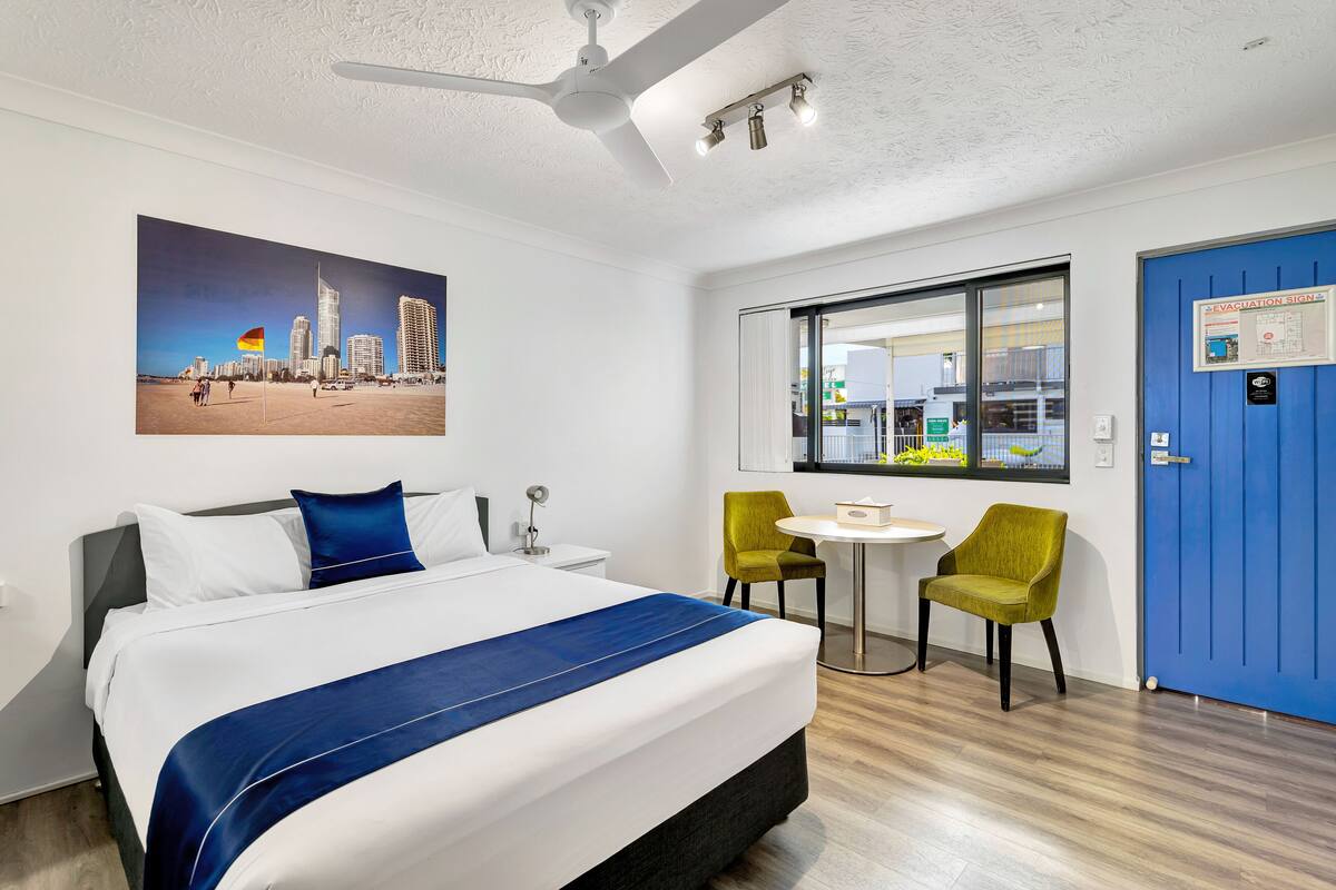 A hotel room with a bed, a desk, two chairs, a window with a view of the city, and a blue door.