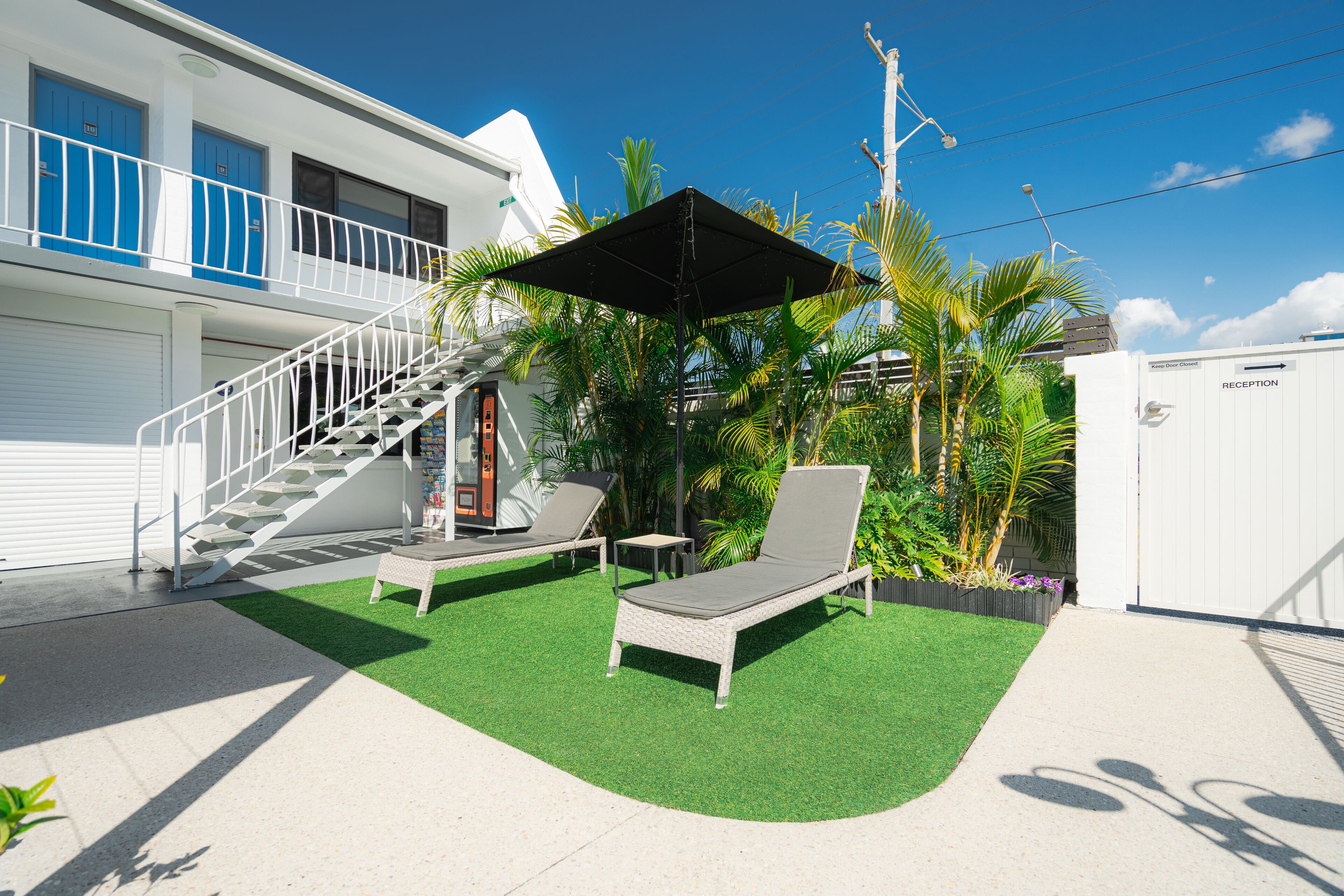 A hotel with a white exterior, a staircase leading to a balcony, two lounge chairs, and a reception area.