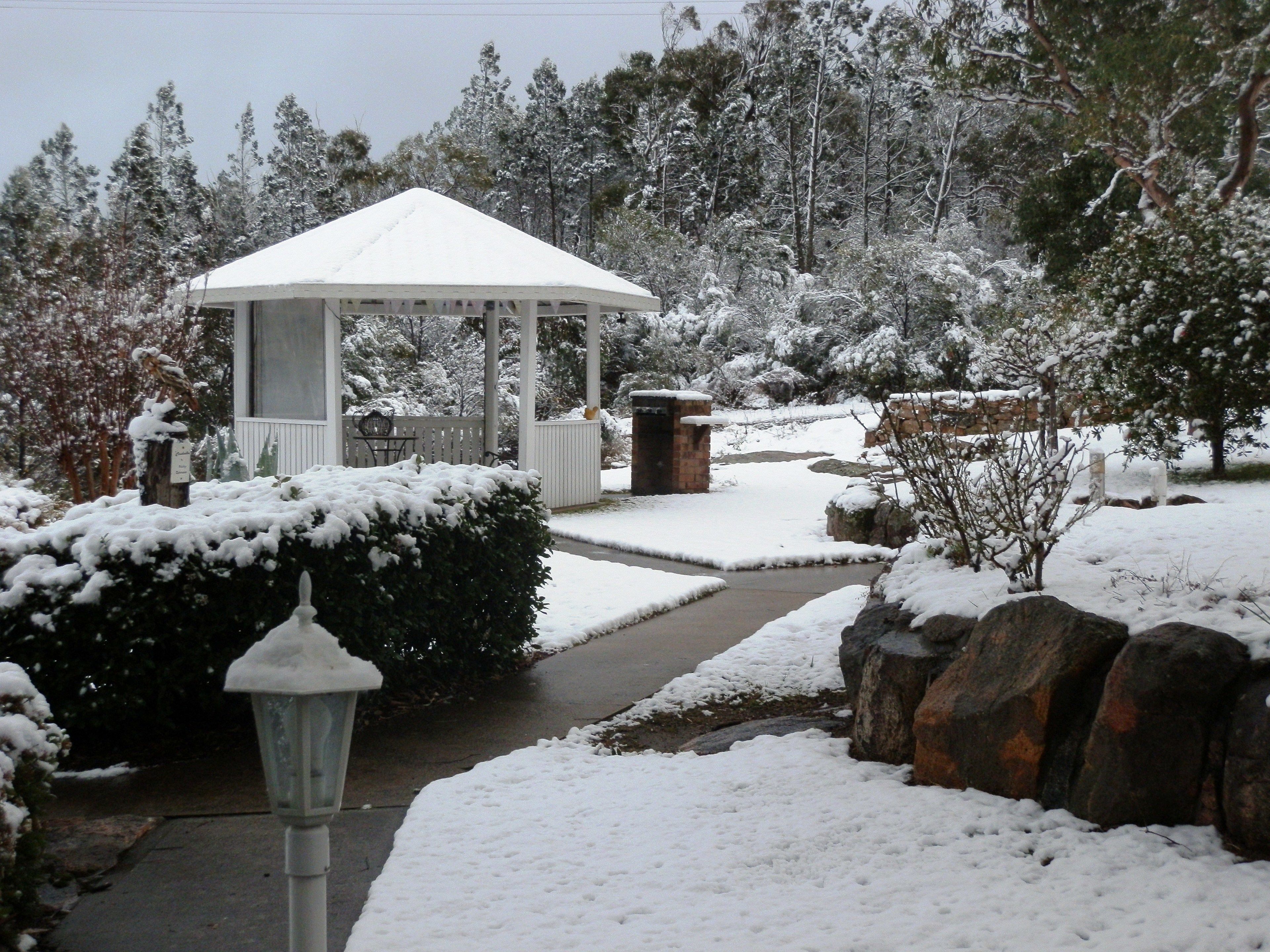 A gazebo with a snow-covered roof, surrounded by snow-laden bushes and trees.