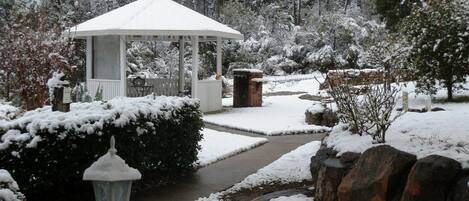 A gazebo with a snow-covered roof, surrounded by snow-laden bushes and trees.