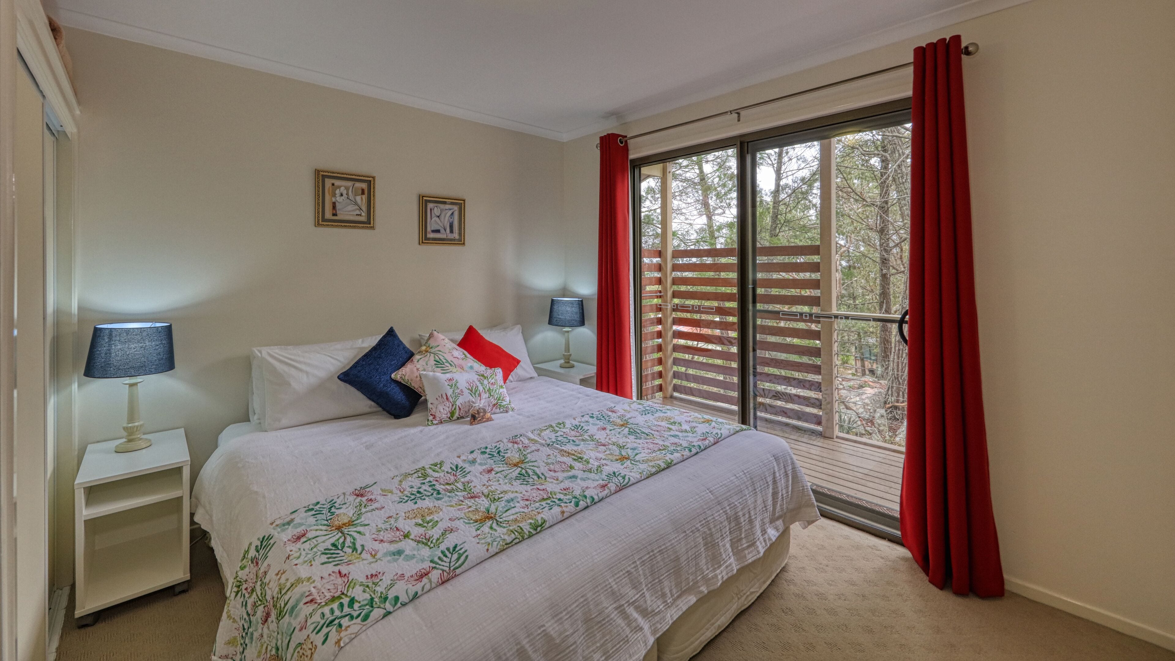 A bedroom with a large bed, two bedside tables with lamps, a window with red curtains, and two framed pictures on the wall.