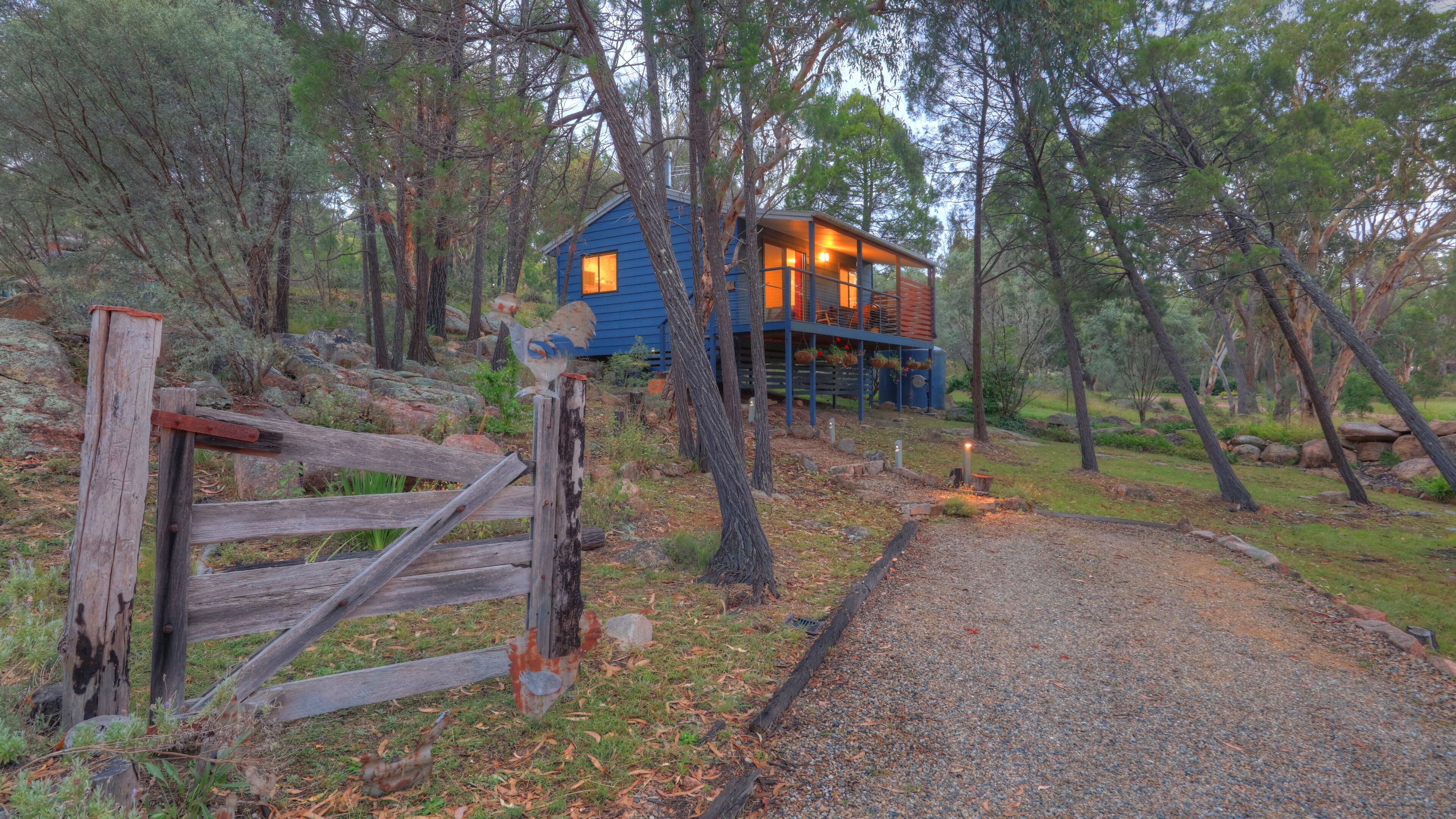 A rustic cabin with a wooden deck, surrounded by trees and a gravel path leading to the entrance.