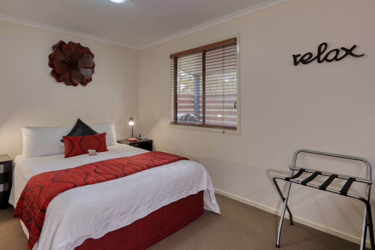 A bedroom with a bed, two bedside tables, a nightstand, a wall decoration, a window with blinds, and a metal chair.