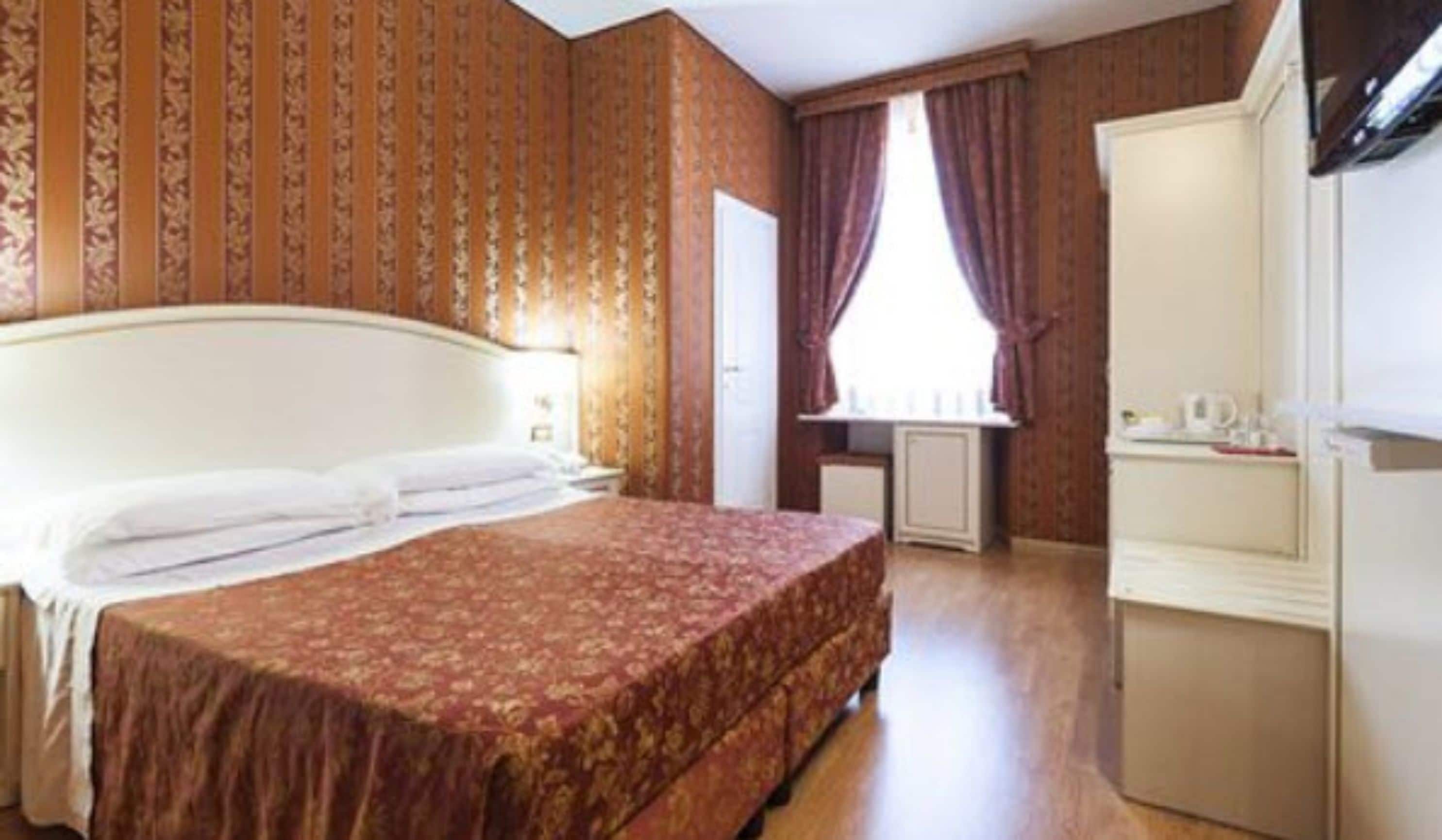 A hotel room with a large bed, a dresser, a window with curtains, and a radiator.