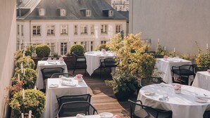 Ein Dachrestaurant mit Tischen für zwei Personen, umgeben von Grünpflanzen und mit Blick auf ein Gebäude.