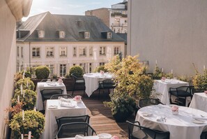 A rooftop dining area with tables set for two, surrounded by greenery and overlooking a building.