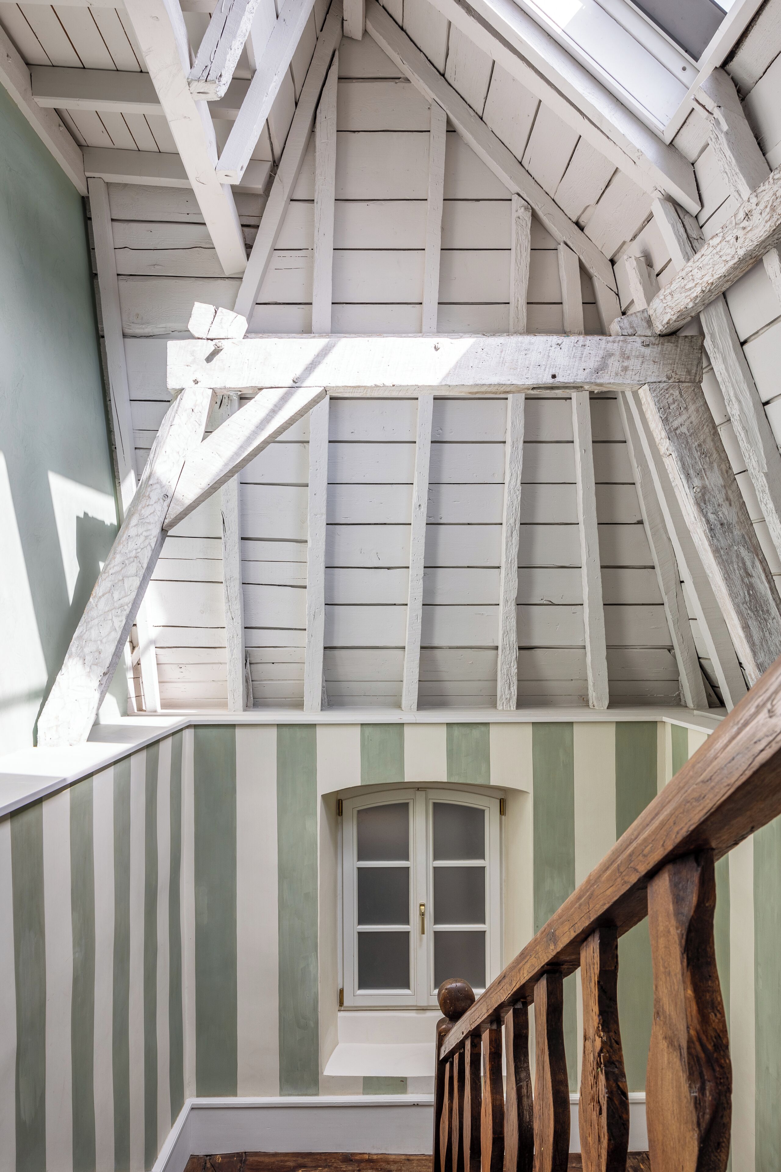Una habitación con techo inclinado, ventana de marco blanco y barandilla de madera.
