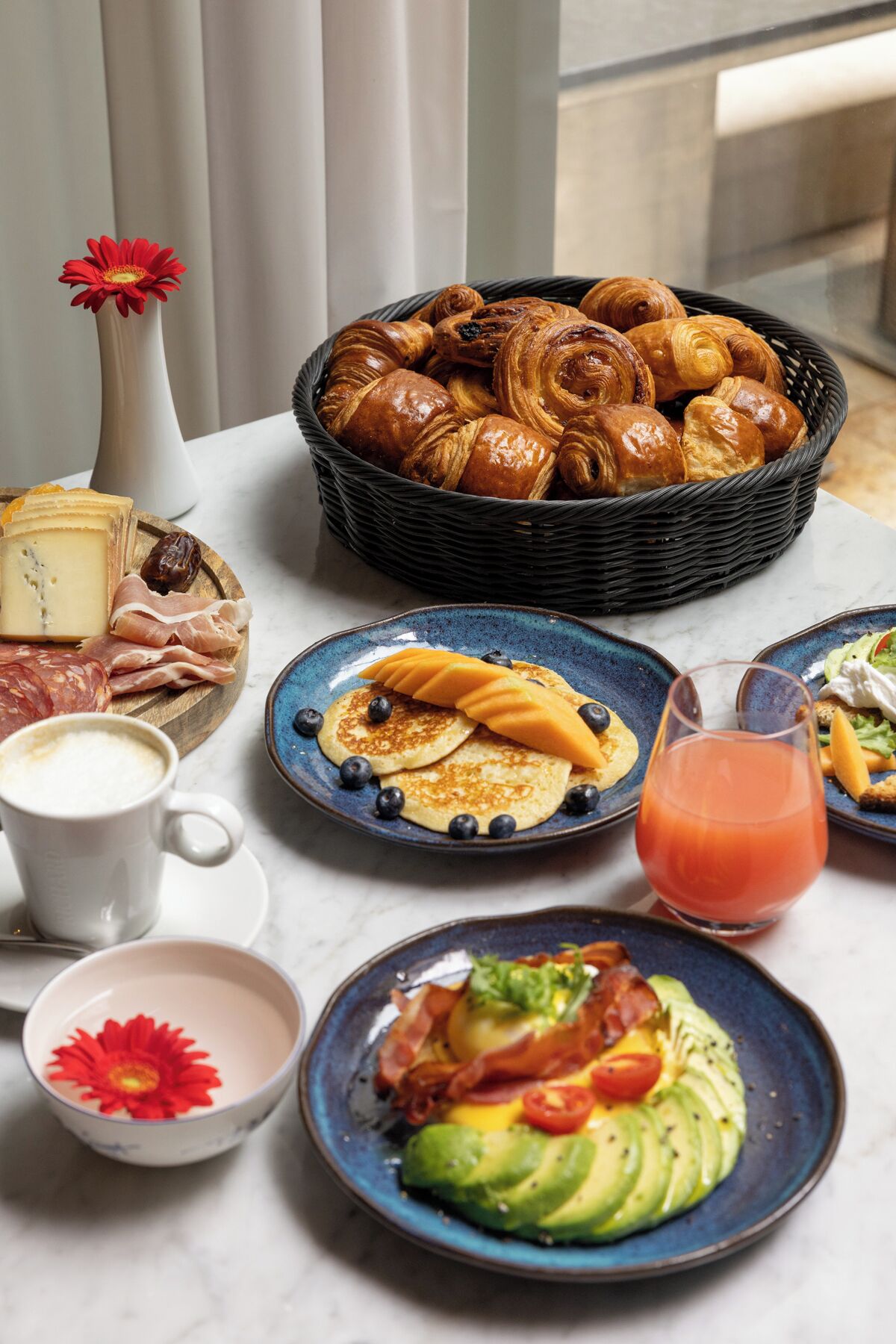 A breakfast table with pastries, cheese, cured meats, pancakes with fruit, a glass of juice, and a bowl of soup.