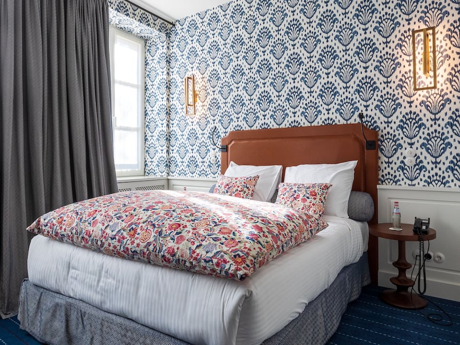 Un dormitorio con una cama grande, papel tapiz estampado, una ventana con cortinas y una mesita de noche con lámpara.