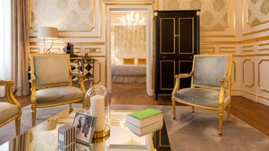 Una habitación lujosa con detalles dorados ornamentales, dos elegantes sillones, una mesa de centro de vidrio con libros y una vela, y una puerta negra grande.