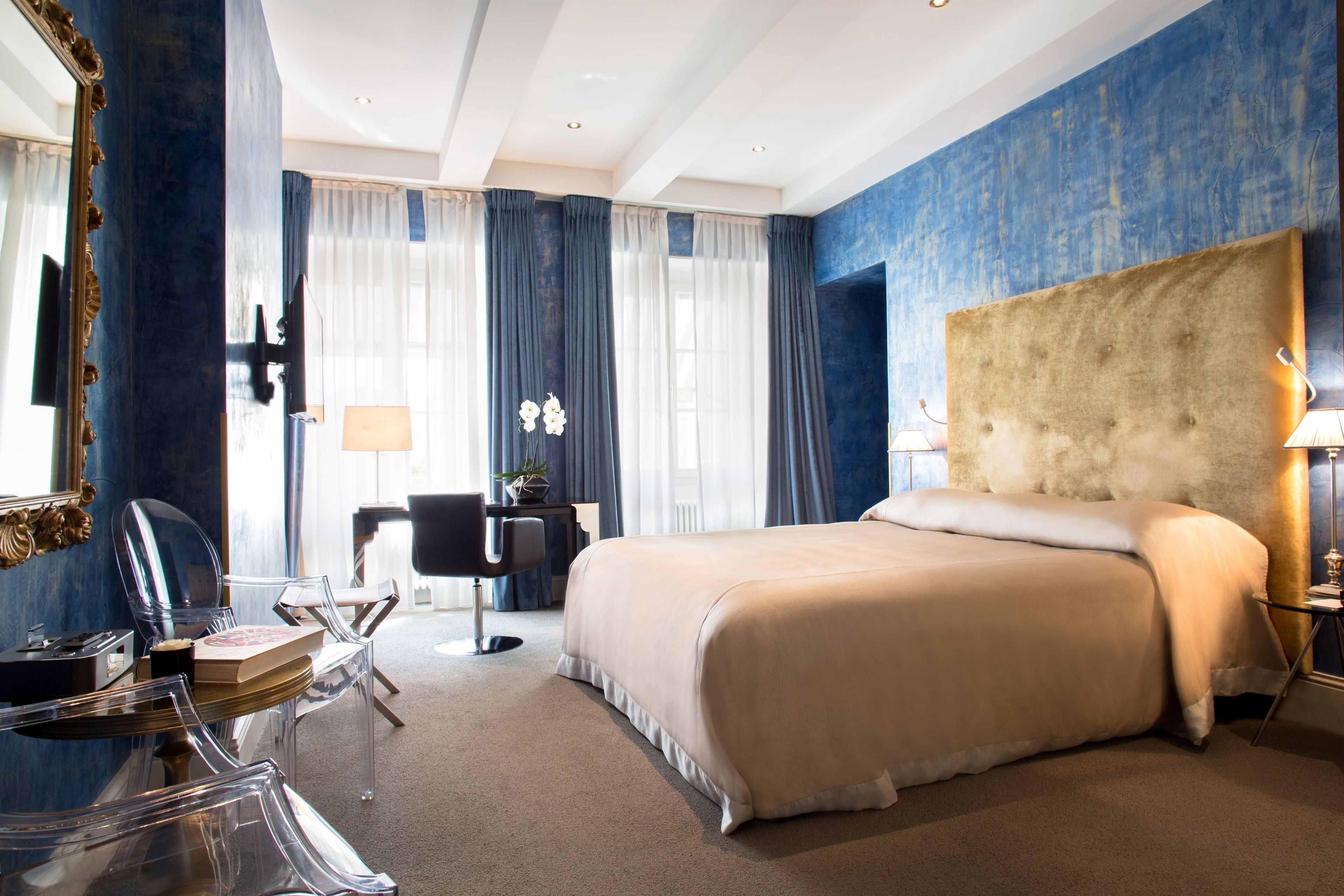 Ein modernes Hotelzimmer mit einem großen Bett, einem Schreibtisch mit Stuhl, einem Spiegel und Blick ins Freie durch große Fenster.
