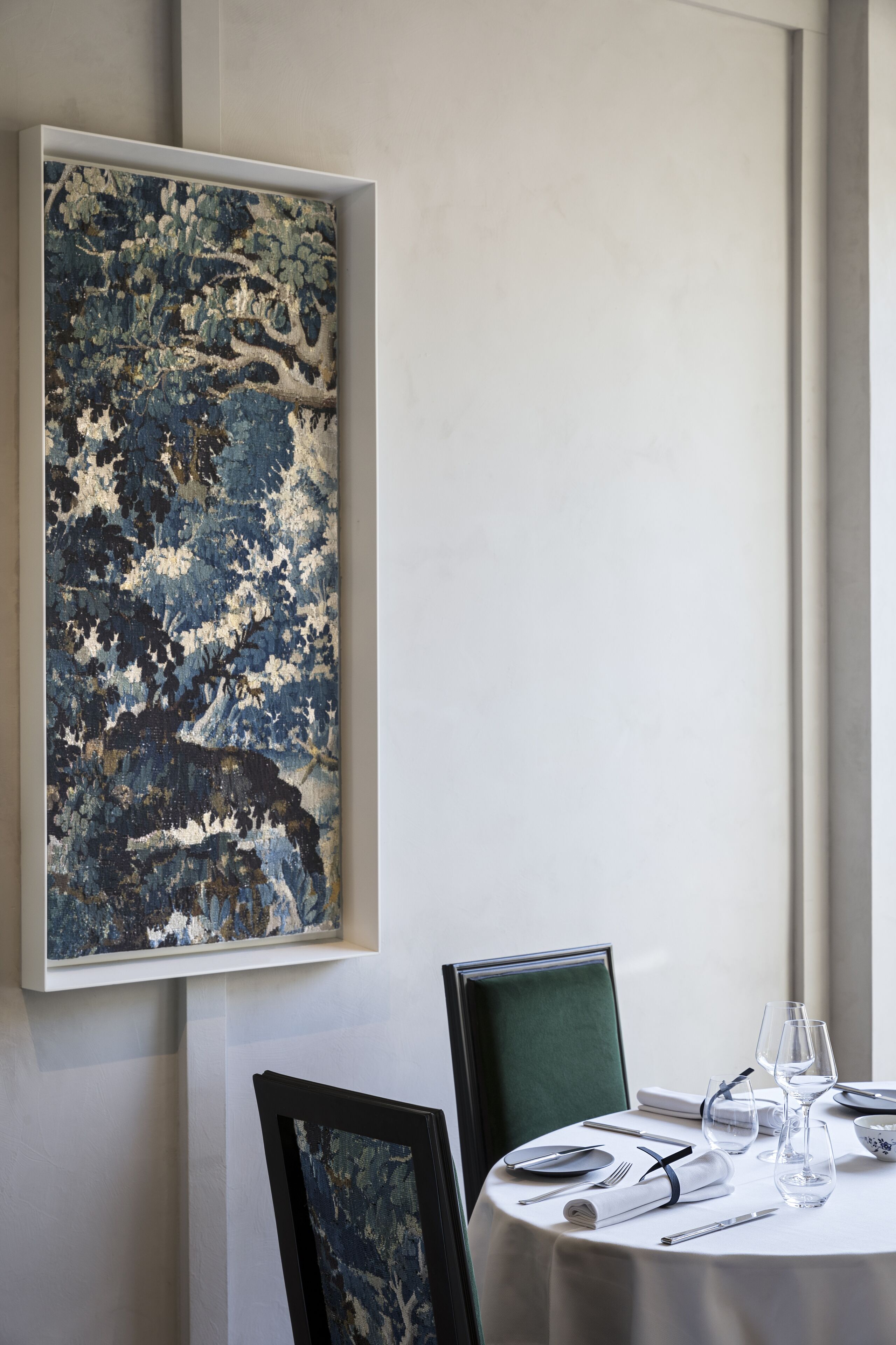 A dining table set with white tablecloth, plates, cutlery, and glasses, with a framed abstract painting above it.