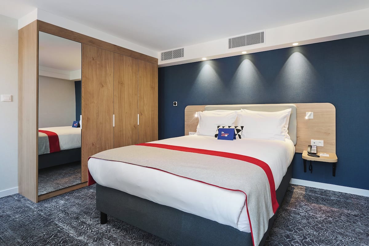 A hotel room with a large bed, a wooden wardrobe, a bedside table with a phone, and a wall-mounted heater.