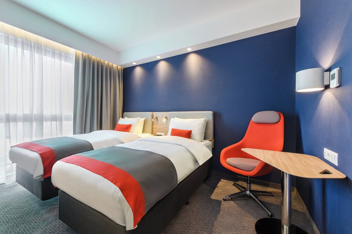 A hotel room with two beds, a desk, a chair, and a wall-mounted lamp.