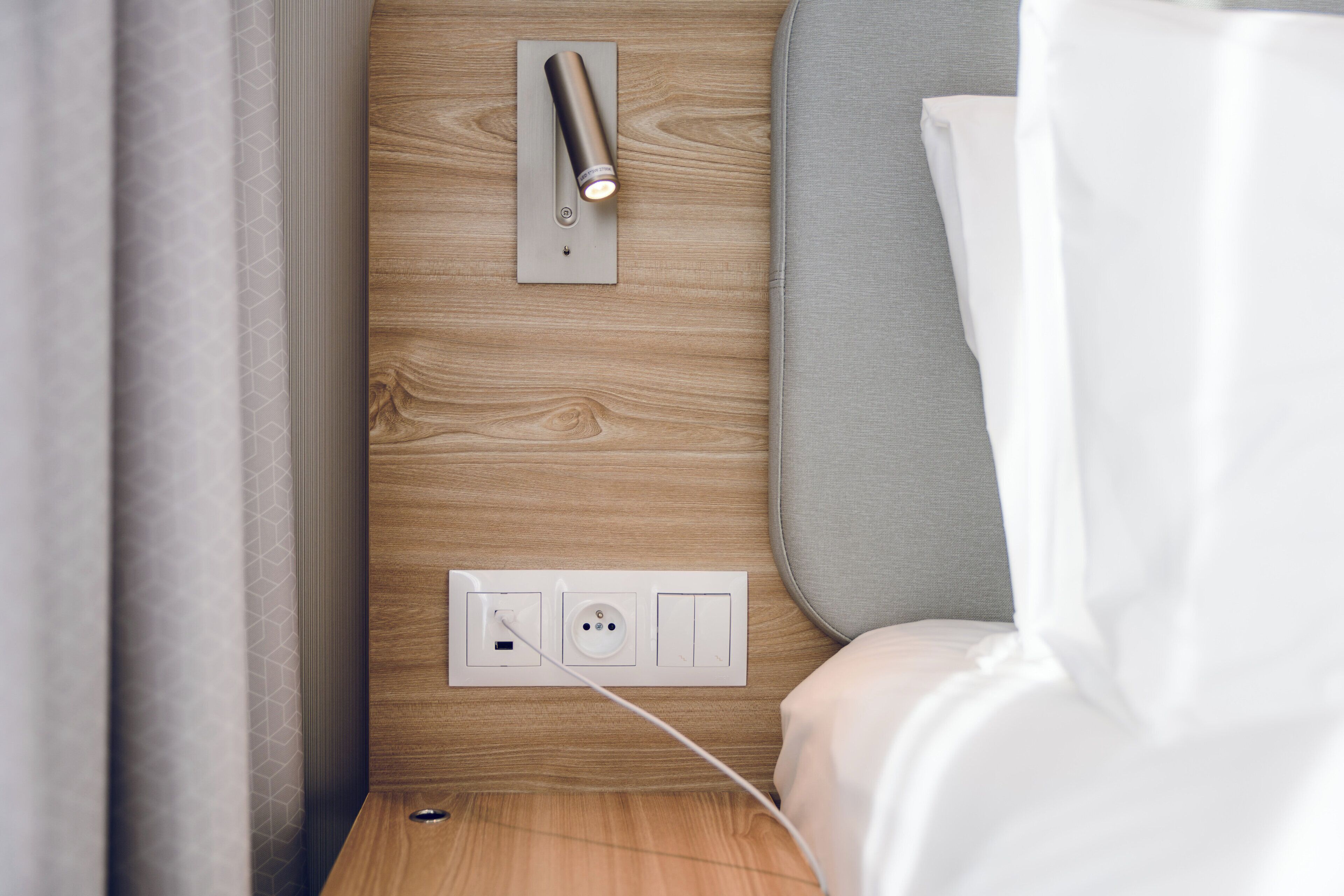 A modern bedroom with a wooden wall, a bedside table, a power outlet, and a lamp.