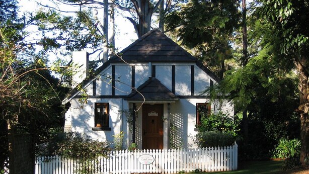 Ein charmantes, eingeschossiges Haus mit weissem Anstrich und dunkler Verzierung, umgeben von einem weissen Zaun und üppiger Bepflanzung.