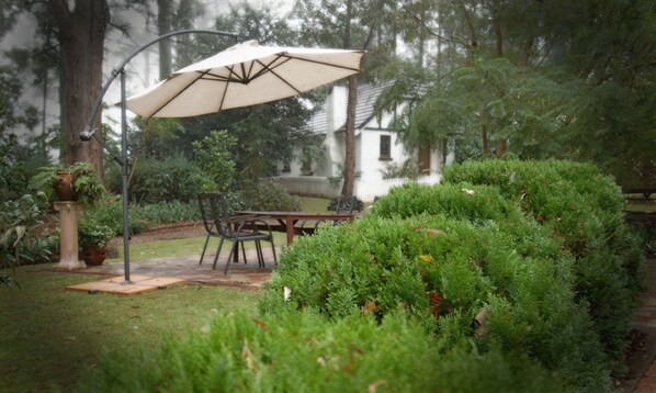 Un patio con mesa y sillas bajo un paraguas grande, rodeado de abundante vegetación y un edificio blanco al fondo.