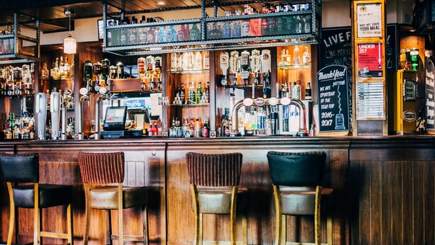 A bar with wooden bar stools, a variety of liquor bottles, and a chalkboard sign. - The Commercial Hotel (Wishaw)