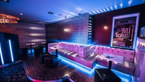A modern lounge area with a neon-lit wall, a seating area with cushioned benches, and a large window displaying a neon sign. - The Commercial Hotel (Wishaw)