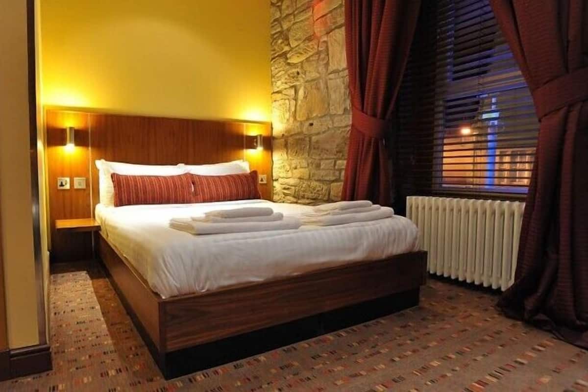 A hotel room with a large bed, wooden headboard, stone wall, and a window with curtains.