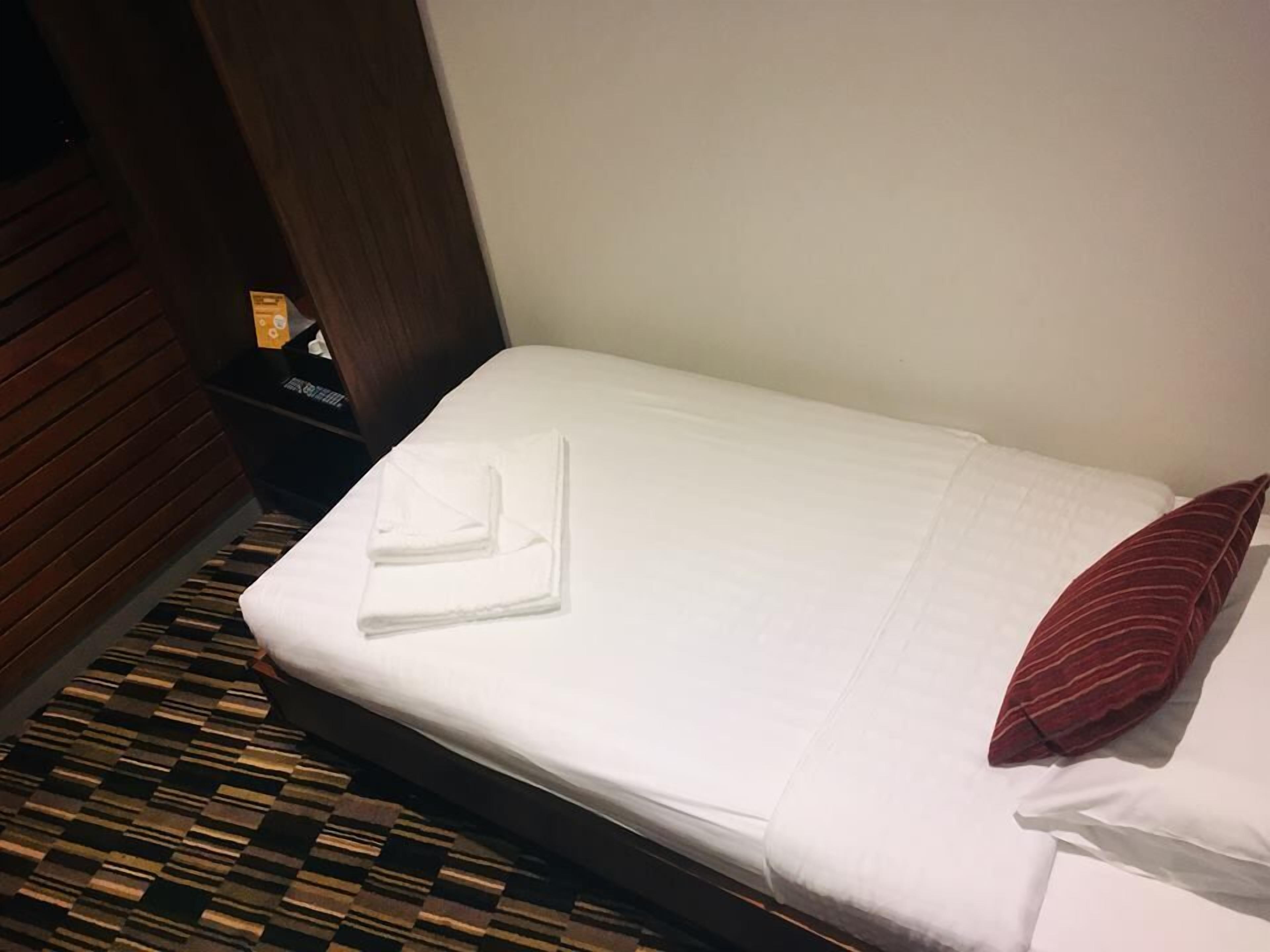 Una cama cuidadosamente tendida con una toalla blanca doblada y un cojín rojo a rayas, una mesita de noche de madera oscura con un objeto pequeño encima, y una alfombra estampada.
