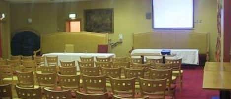 Une salle de conférence avec des rangées de chaises en bois, un écran de projection, un lit et une petite table.