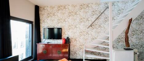 Une pièce moderne avec une grande fenêtre, un téléviseur à écran plat posé sur un meuble en bois, un escalier blanc et un radiateur.