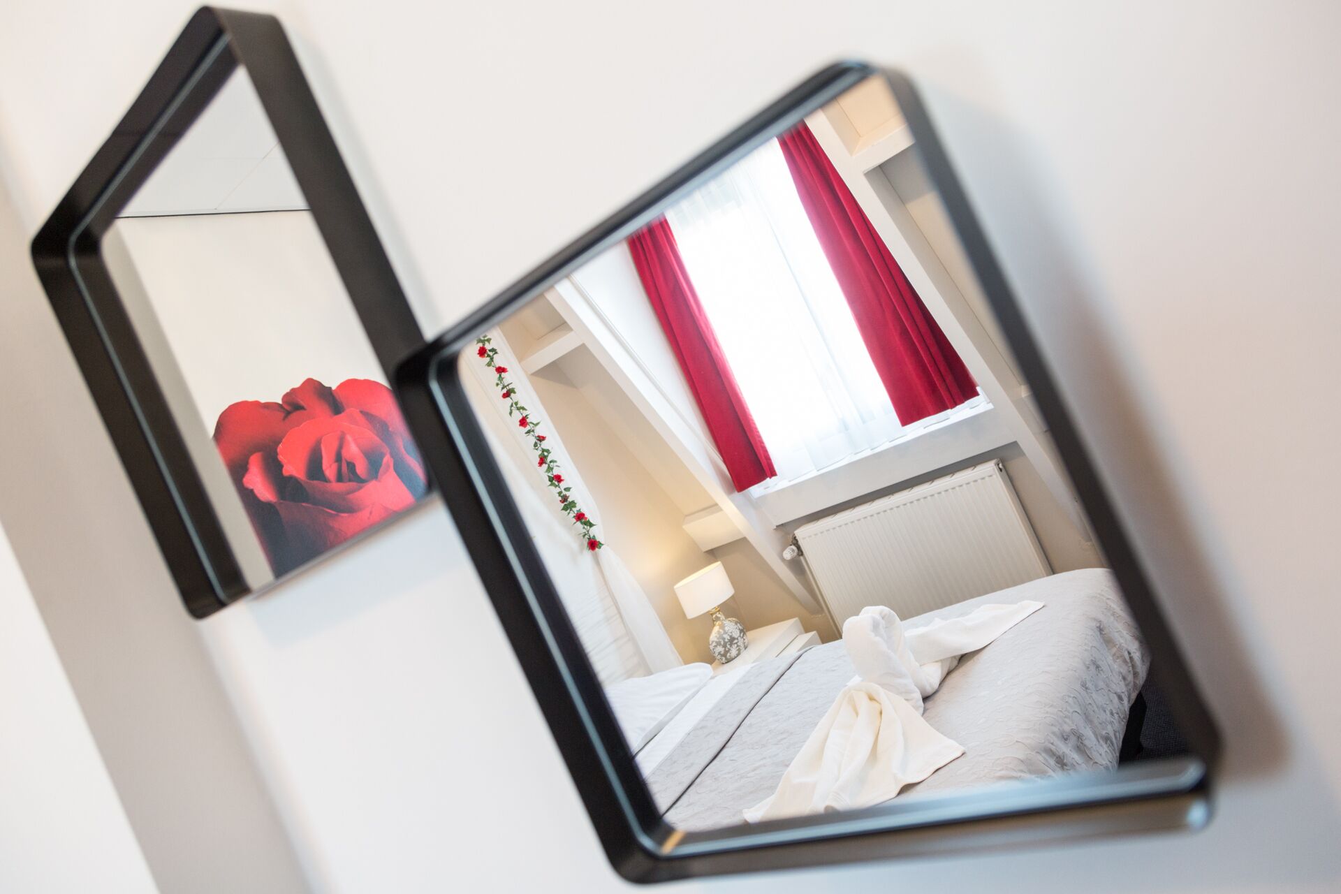 Un miroir reflétant une pièce avec un lit, une fenêtre ornée de rideaux rouges et un tableau représentant une rose accroché au mur.
