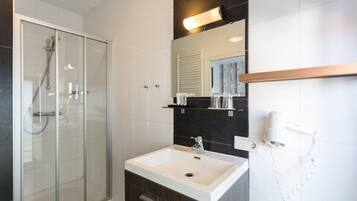 Une salle de bain moderne avec une douche à parois de verre, un lavabo blanc et un miroir surmonté d’une étagère en bois.