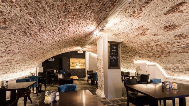 Um café subterrâneo com paredes de pedra, mesas e cadeiras.