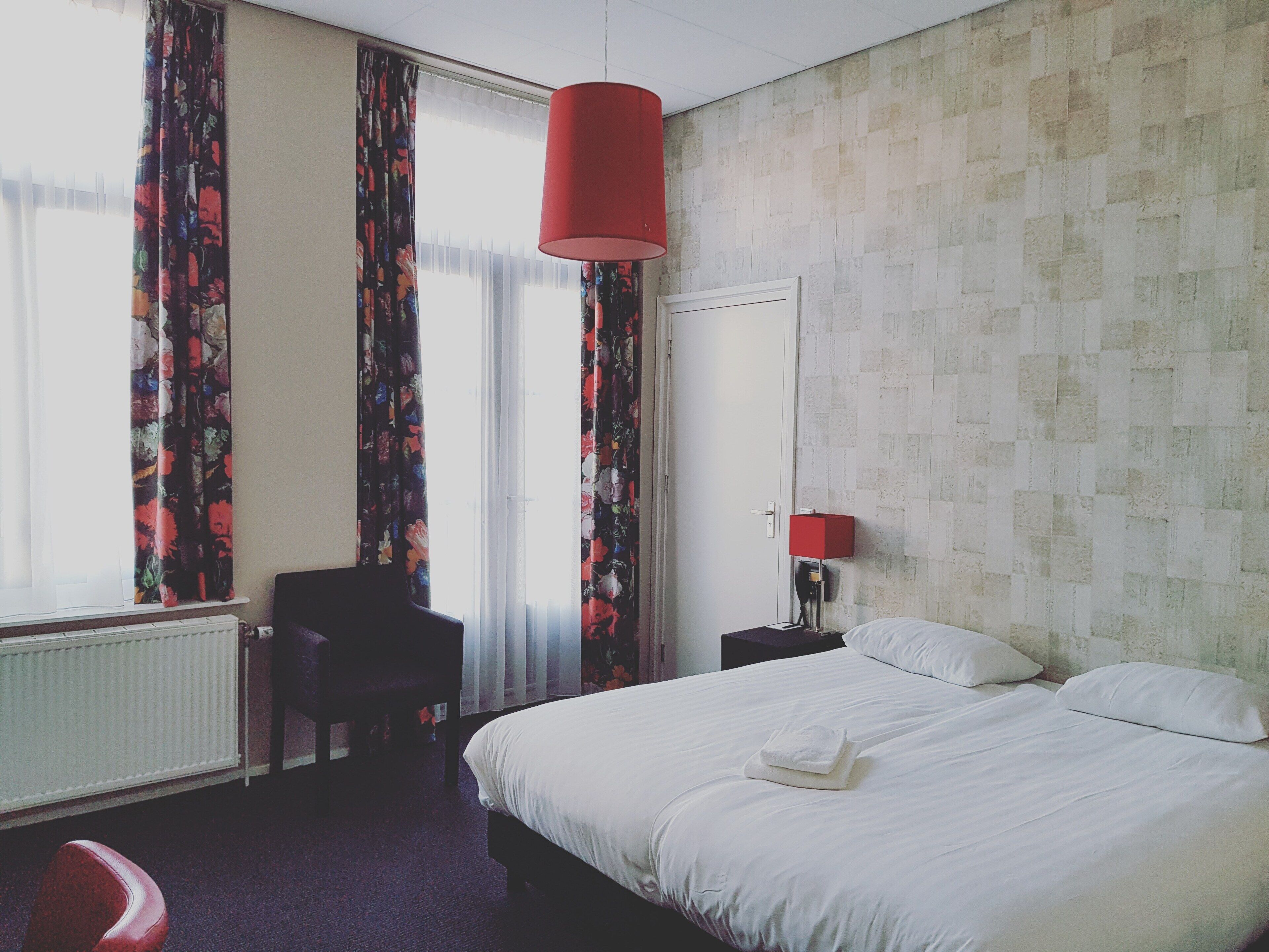 Ein Hotelzimmer mit einem Bett, einem Sessel, einer roten Lampe und einem Fenster mit blumigen Vorhängen.