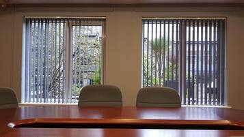 A conference room with a long table and four chairs, large windows with vertical blinds, and a view of trees and buildings outside.