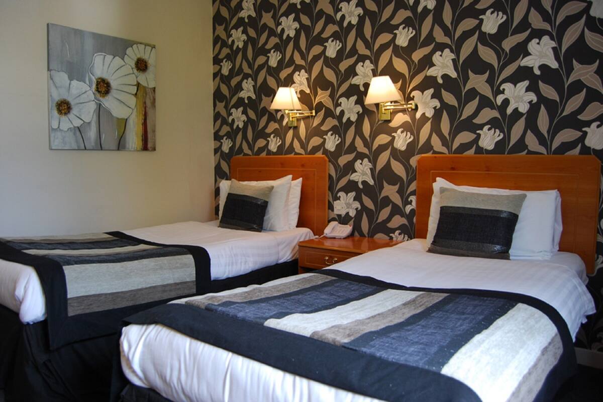 A hotel room with two beds, a floral wallpaper, a painting of flowers, and two wall-mounted lamps.