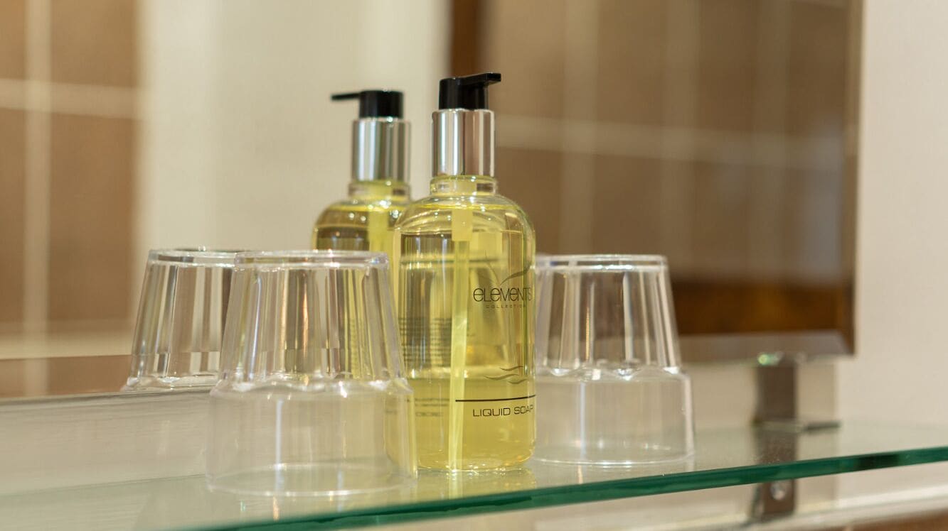 Un baño con un estante de vidrio que contiene dos botellas de jabón líquido y varios vasos de cristal.