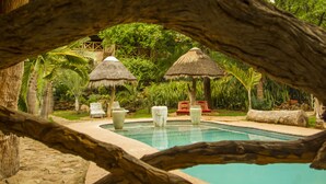 Un espace piscine agrémenté de parasols en chaume, de chaises longues et d’un jardin tropical en arrière-plan.