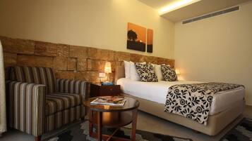 A hotel room with a stone headboard, a bed with patterned bedding, a striped armchair, a small round table with a magazine, and two wall-mounted lamps.