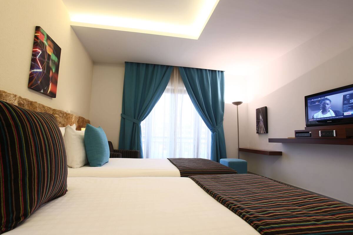 A hotel room with two beds, a television, a painting, and a window with curtains.