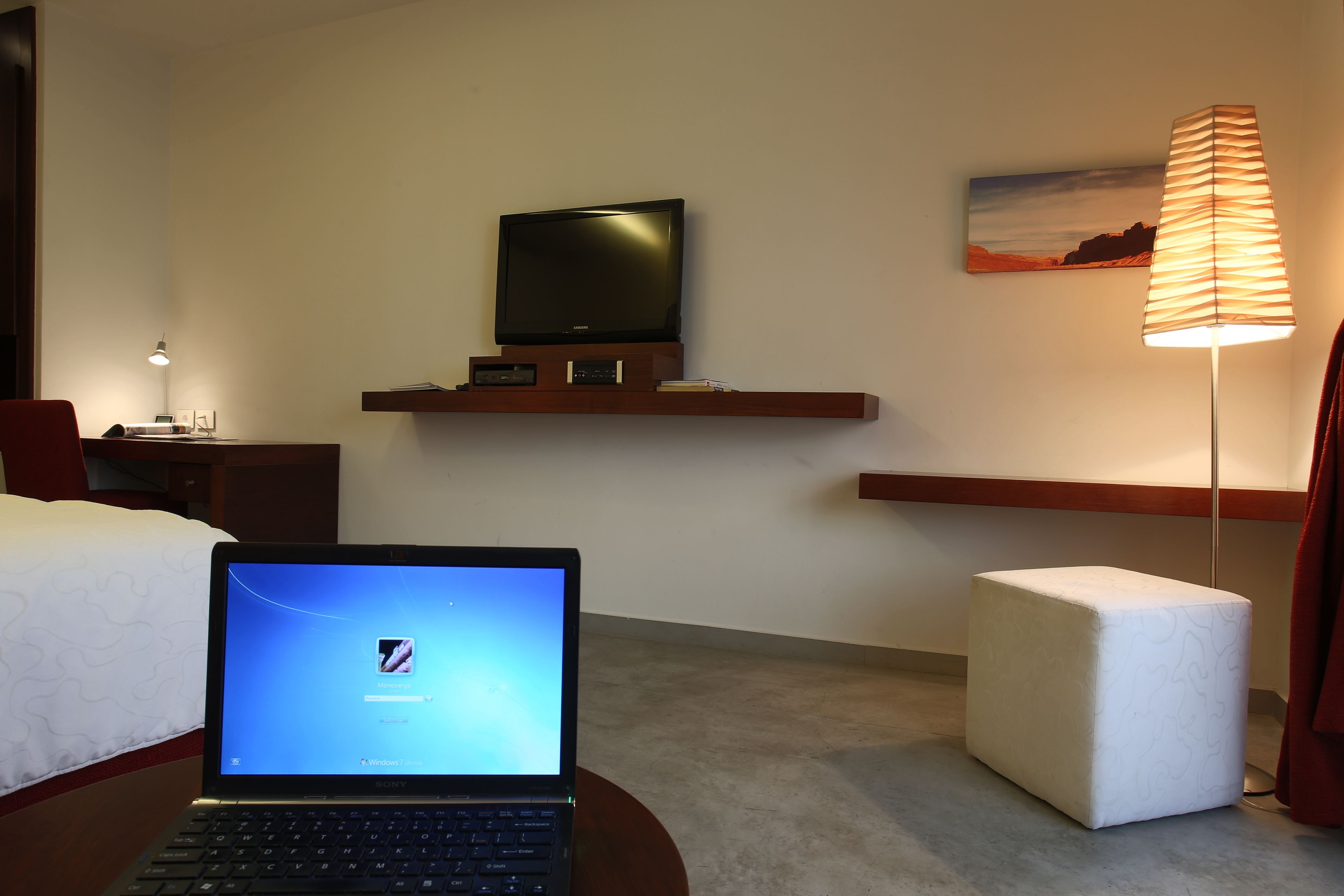 A hotel room with a bed, a desk with a laptop, a TV on a shelf, a lamp, and a small ottoman.