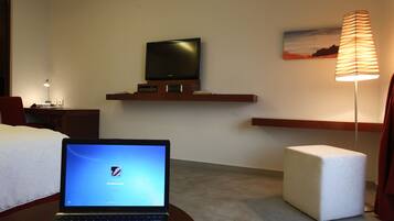 A hotel room with a bed, a desk with a laptop, a TV on a shelf, a lamp, and a small ottoman.