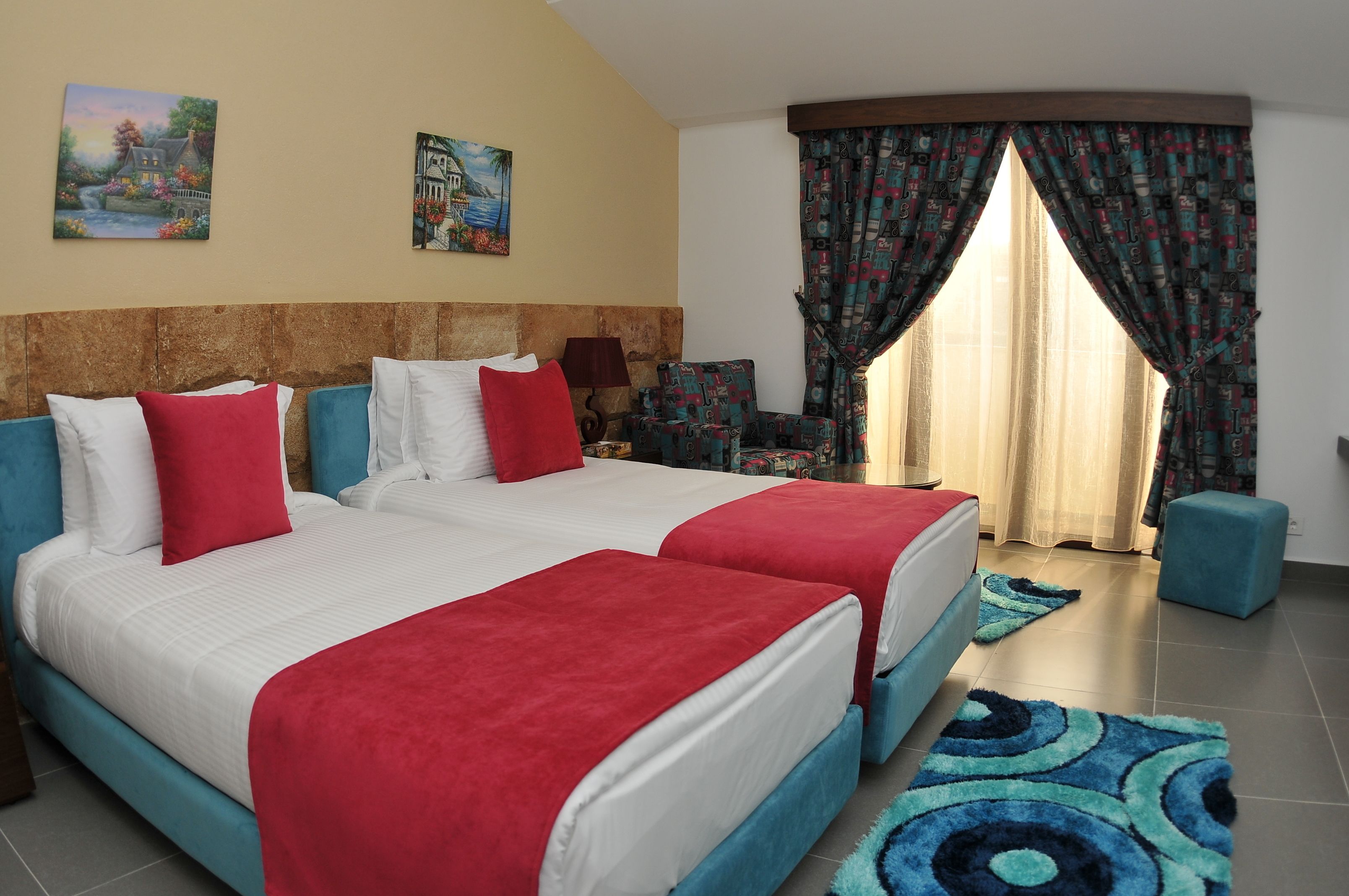 A hotel room with two beds, a sofa, a chair, and a small table. There are two paintings on the wall and a window with curtains.