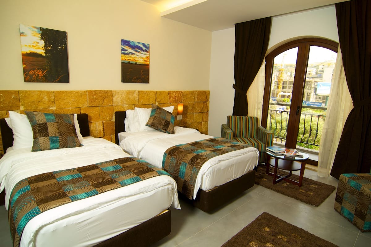 A hotel room with two beds, a small table, and a view of the city through a large window.