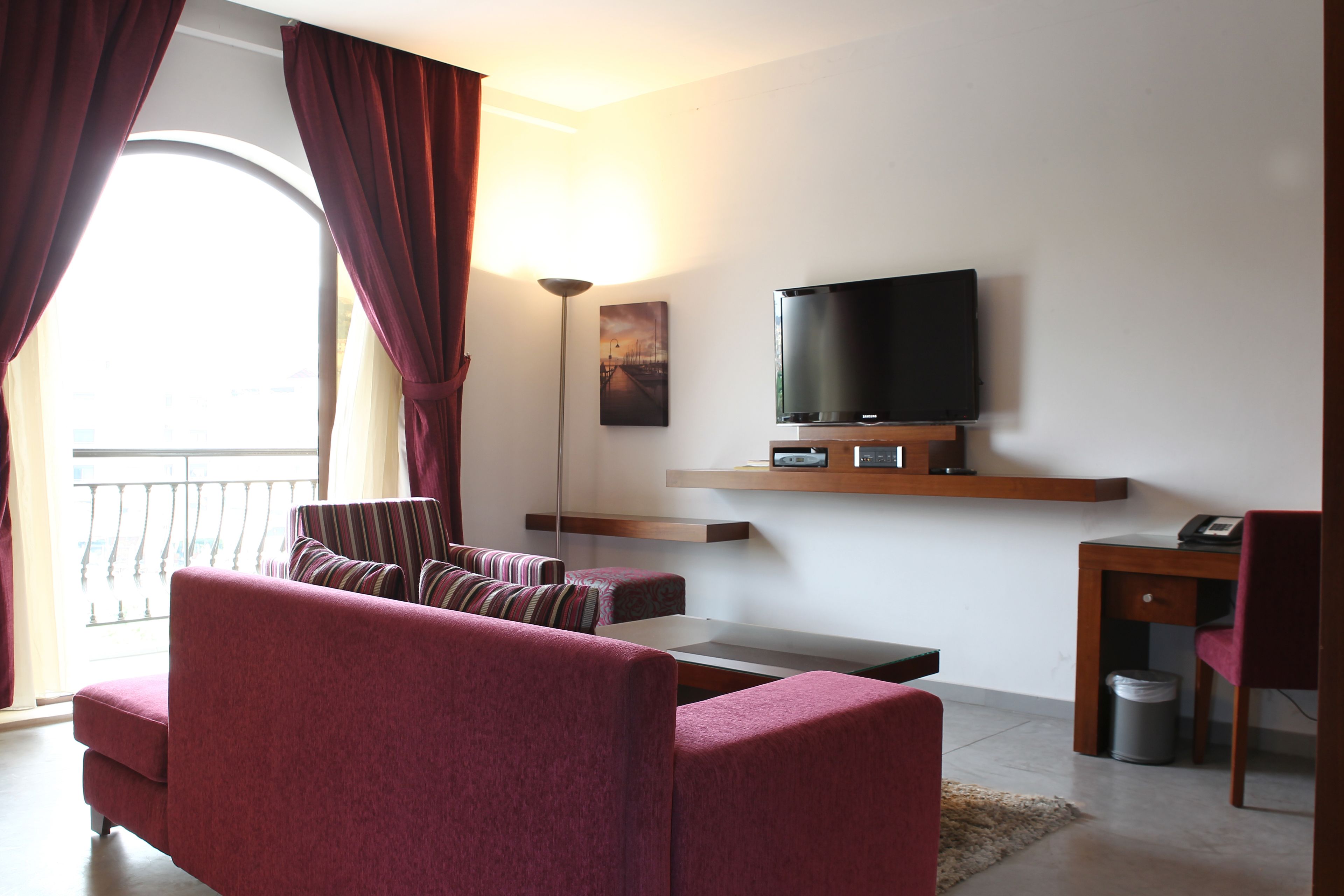 A modern living room with a sofa, a coffee table, a TV on a wooden shelf, and a balcony with curtains.