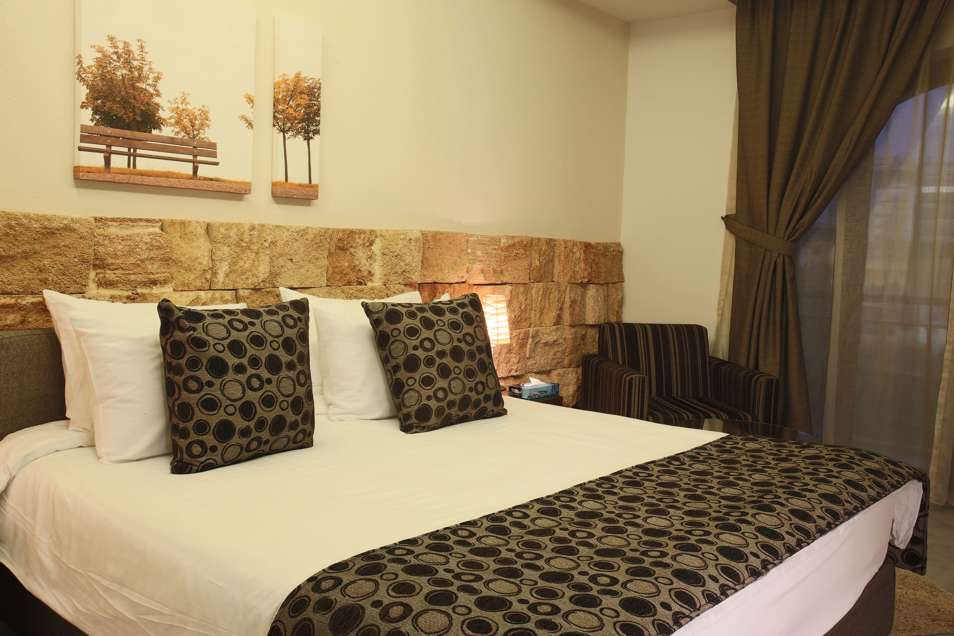 Habitación de hotel con cabecera de piedra, una cama grande con ropa de cama estampada, dos cuadros colgados en la pared y vistas al exterior a través de una ventana con cortinas.